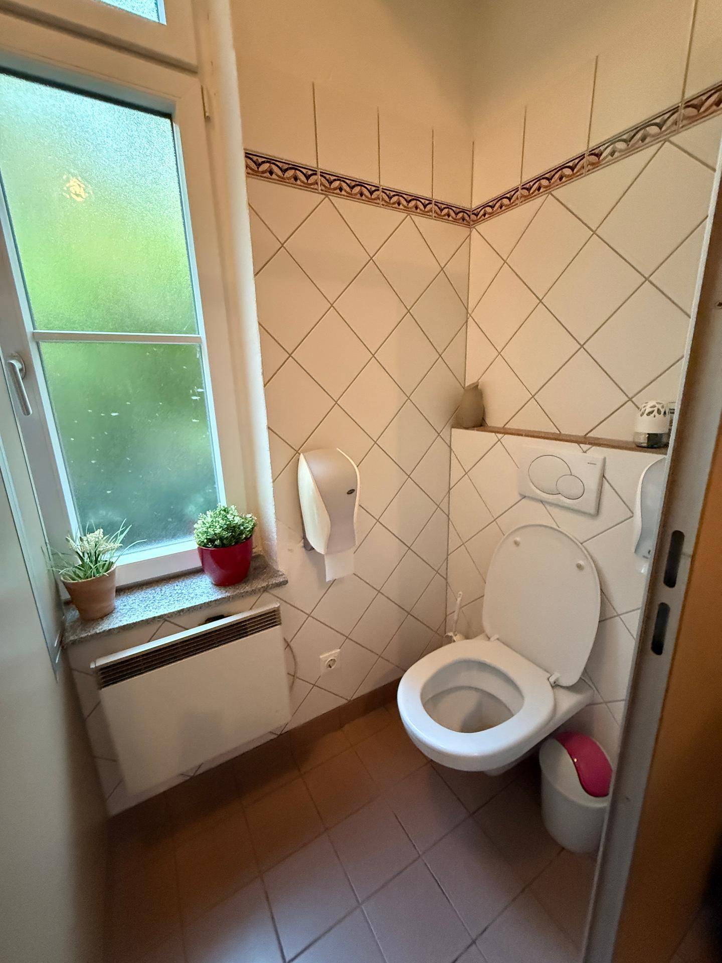Toilettenkabine mit Fenster, WC und einem kleinen Heizkörper, ausgestattet mit hellen Wandfliesen.