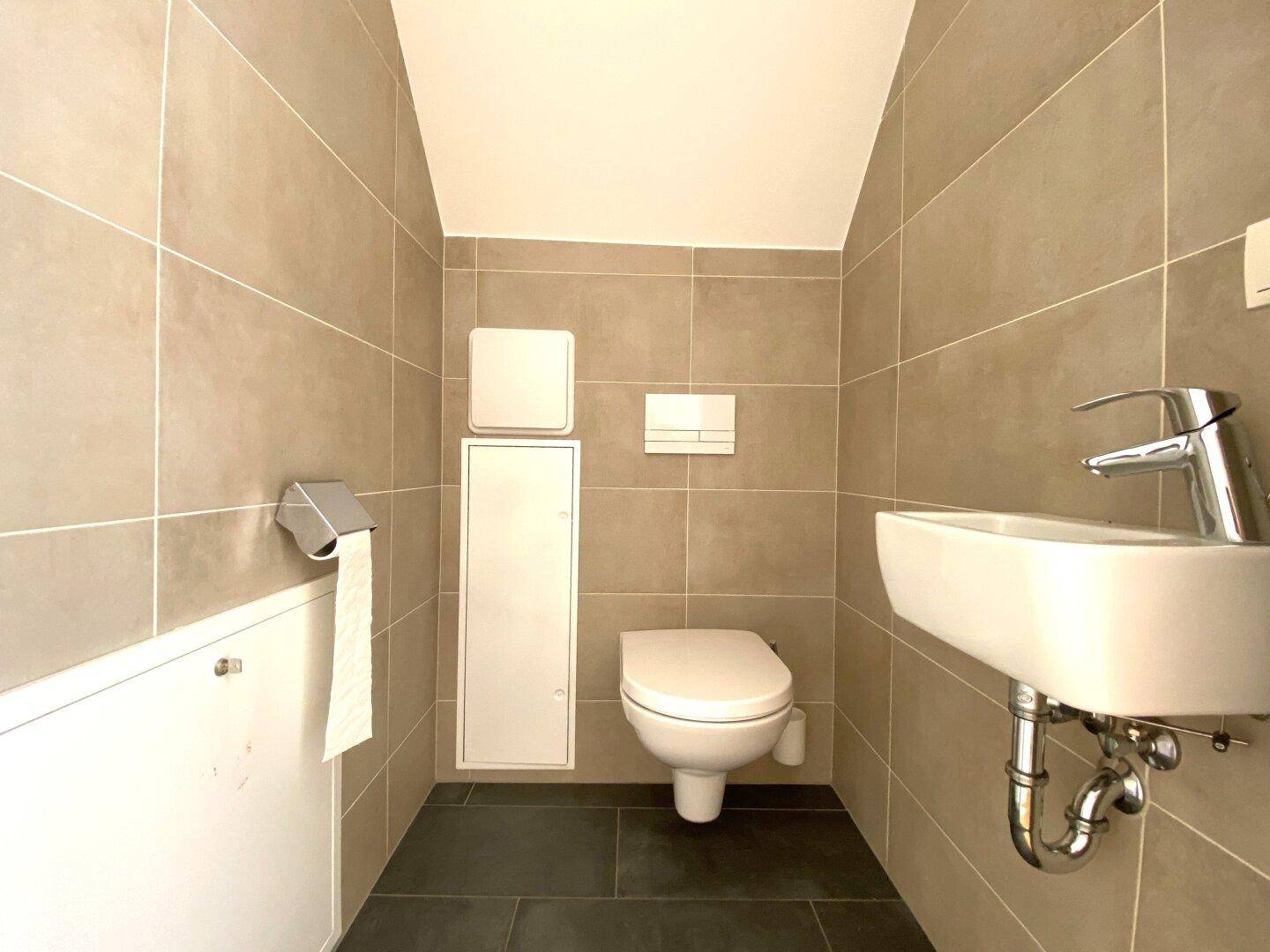 Gäste-WC mit beigefarbenen Wandfliesen, wandhängendem WC und kleinem Handwaschbecken.