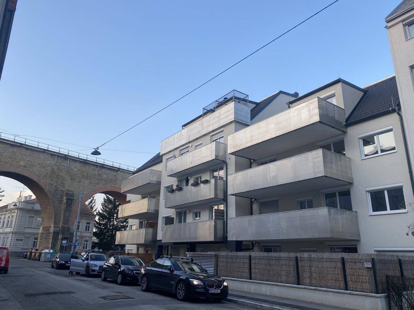 Modernes Mehrfamilienhaus mit mehreren Balkonen und einer historischen Brücke im Hintergrund.