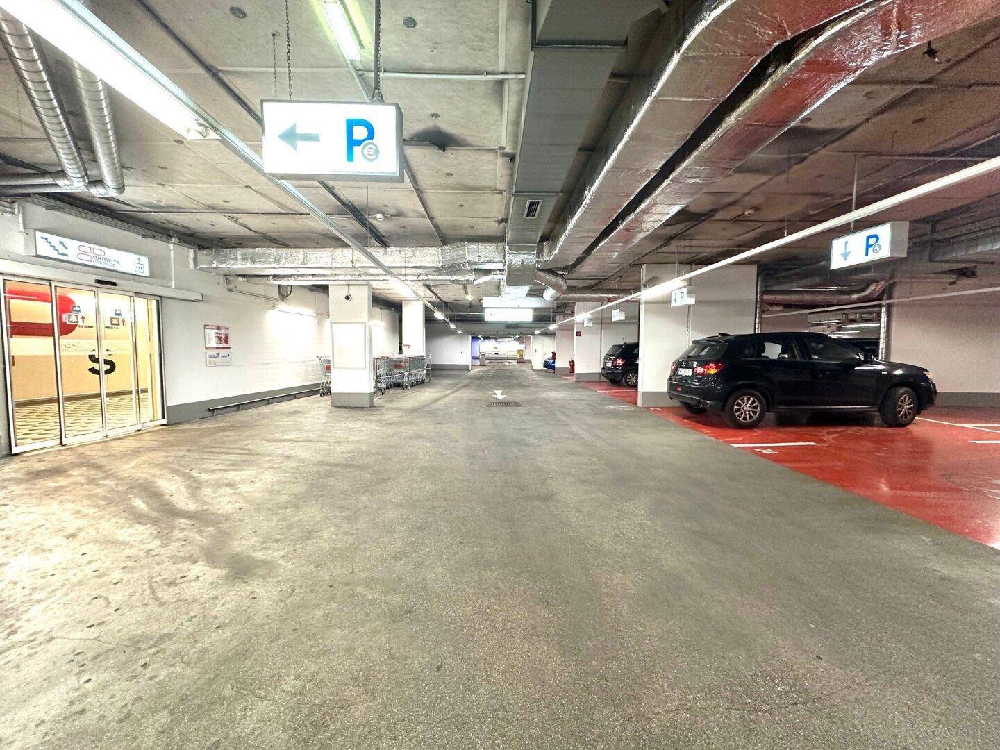 Geräumige Tiefgarage mit Betonboden, sichtbaren Leitungen und geparkten Autos, gut beleuchtet.