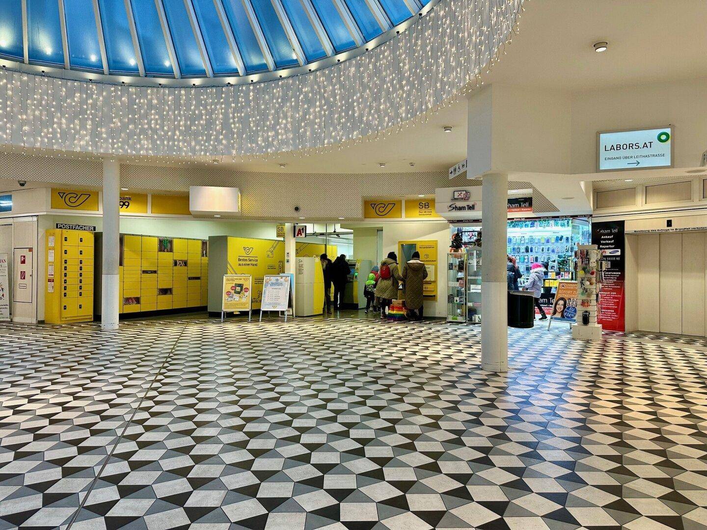 Postfiliale in der Einkaufspassage mit gelben Schließfächern und einem runden Glasdach, das für Helligkeit sorgt.