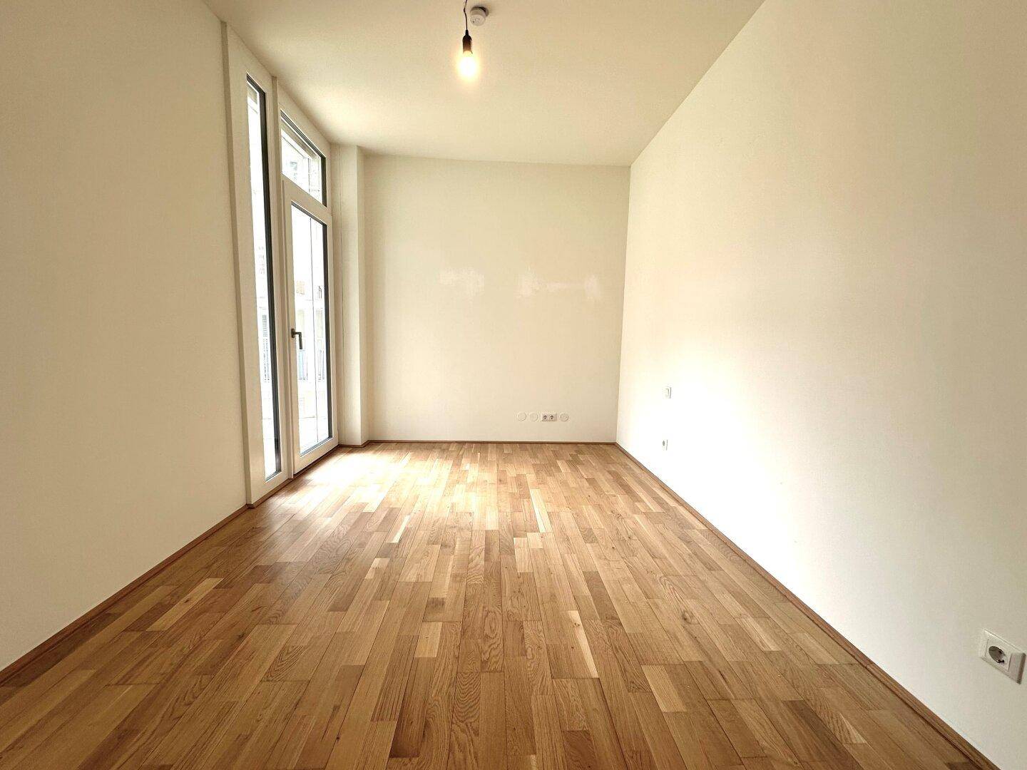 Leeres Zimmer mit hellem Holzfußboden und einer Fenstertür, ideal als Schlafzimmer oder Arbeitszimmer.