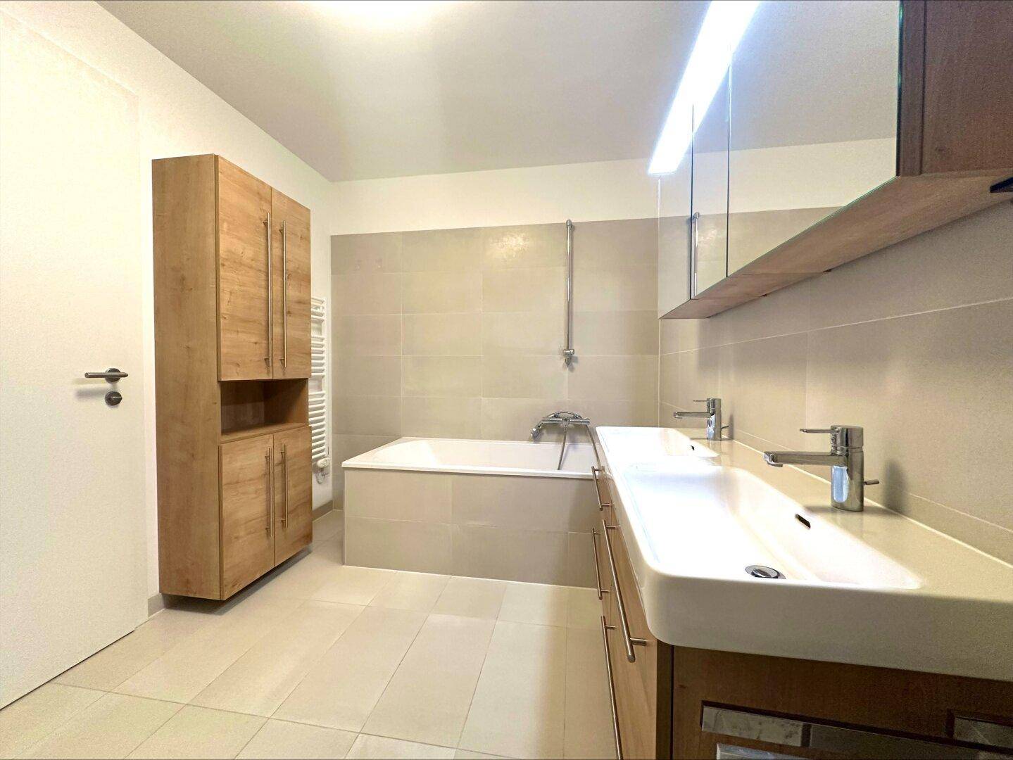 Modernes Badezimmer mit Doppelwaschbecken, Badewanne und hellen Fliesen, ausgestattet mit Handtuchheizkörper.