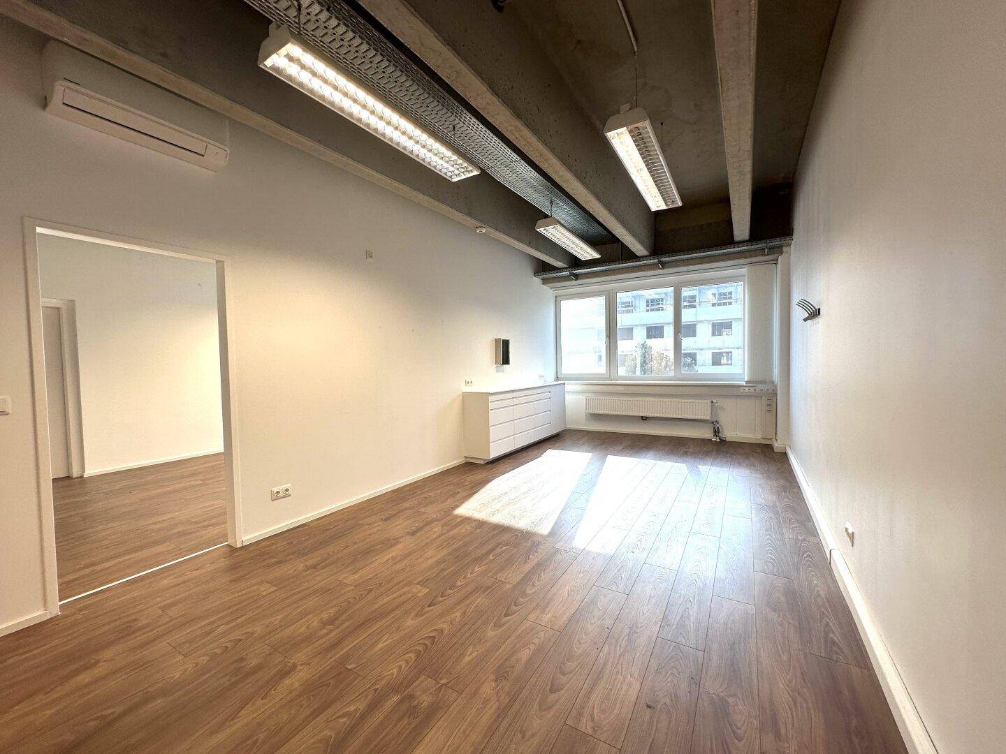Helles Büro mit Küchenzeile, großen Fenstern und Holzboden unter einer Sichtbetondecke.