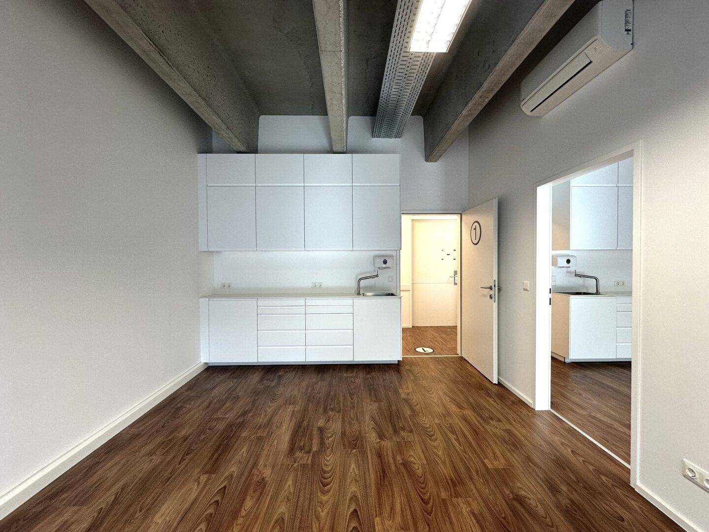 Bürofläche mit Küchenzeile, Holzboden und industriellem Charme durch sichtbare Deckenstrukturen.