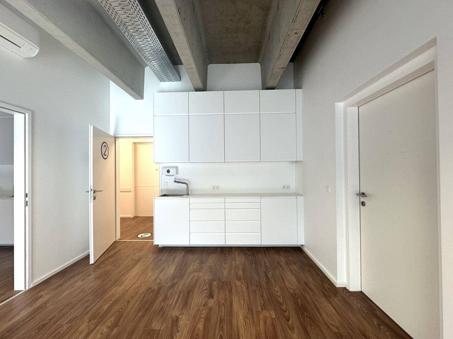 Modernes Büro mit integrierter Küchenzeile, Holzboden und sichtbaren Deckenbalken, offen gestaltet.