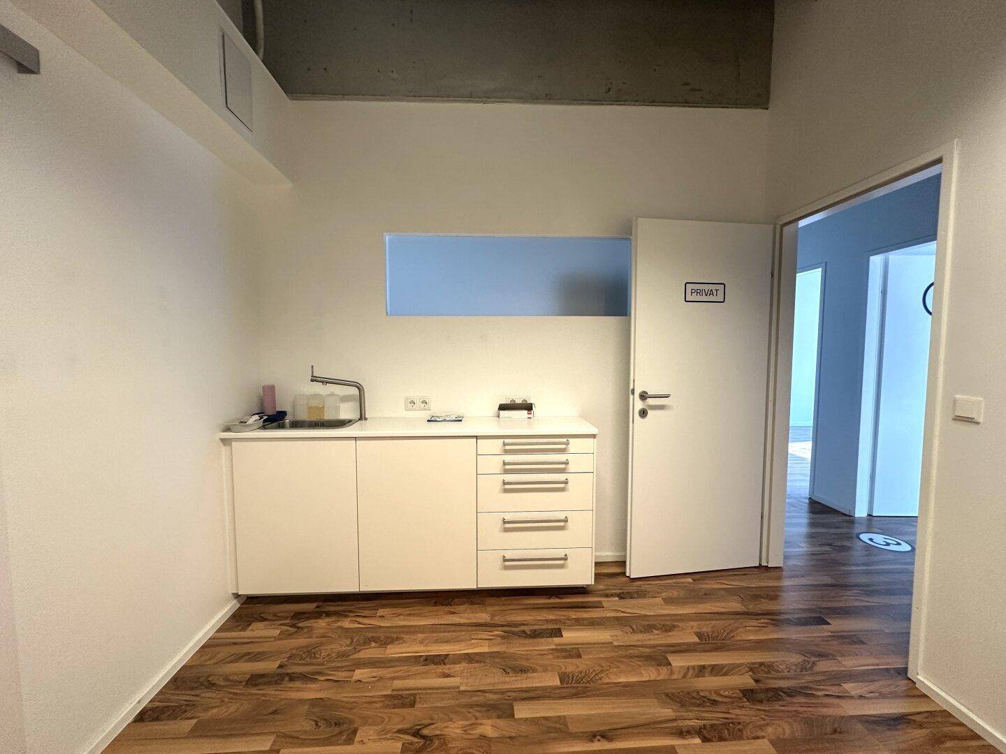 Kleiner Bürobereich mit Küchenzeile, Holzboden und einer Tür zum Privatbereich, gut beleuchtet.
