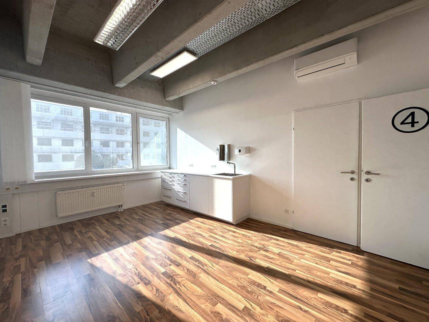 Geräumiges Büro mit Küchenzeile, großen Fenstern und Klimaanlage unter einer Sichtbetondecke.
