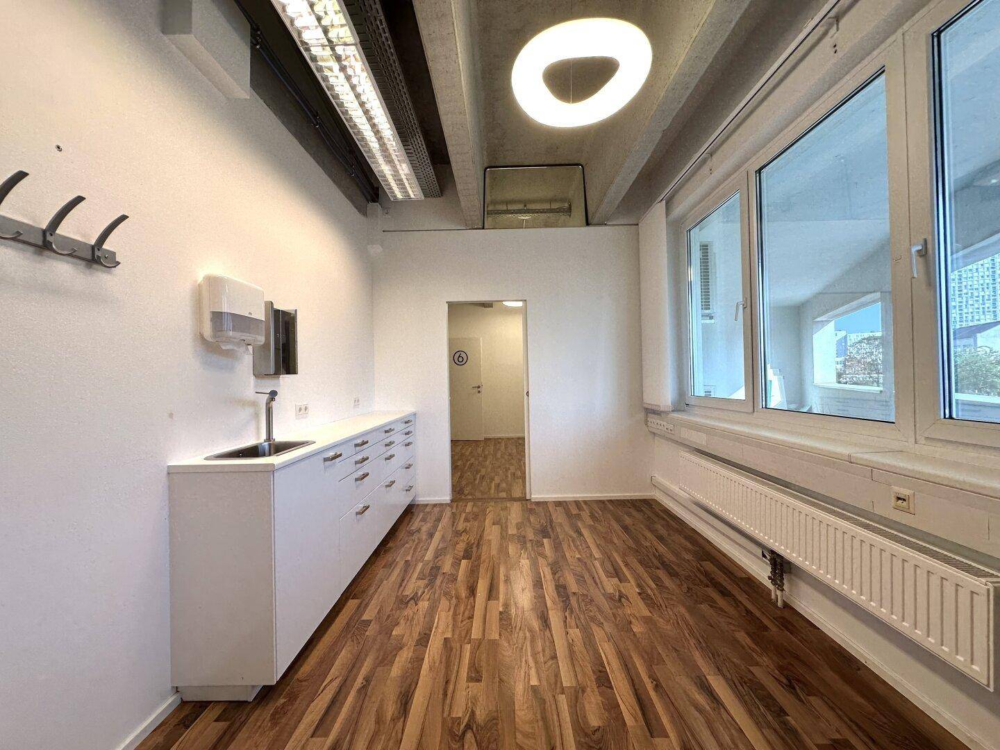Büro mit Küchenzeile, großen Fenstern, Holzboden und Garderobe unter Sichtbetondecke.