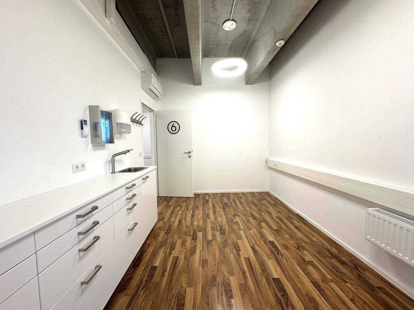 Modernes Büro mit Küchenzeile, Holzboden und Garderobe, beleuchtet von runden Deckenleuchten.