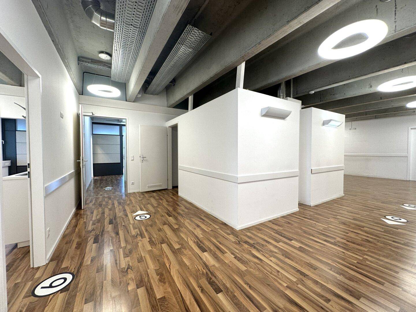 Offener Bürobereich mit Holzboden, modernen Deckenleuchten und sichtbaren Betonelementen.