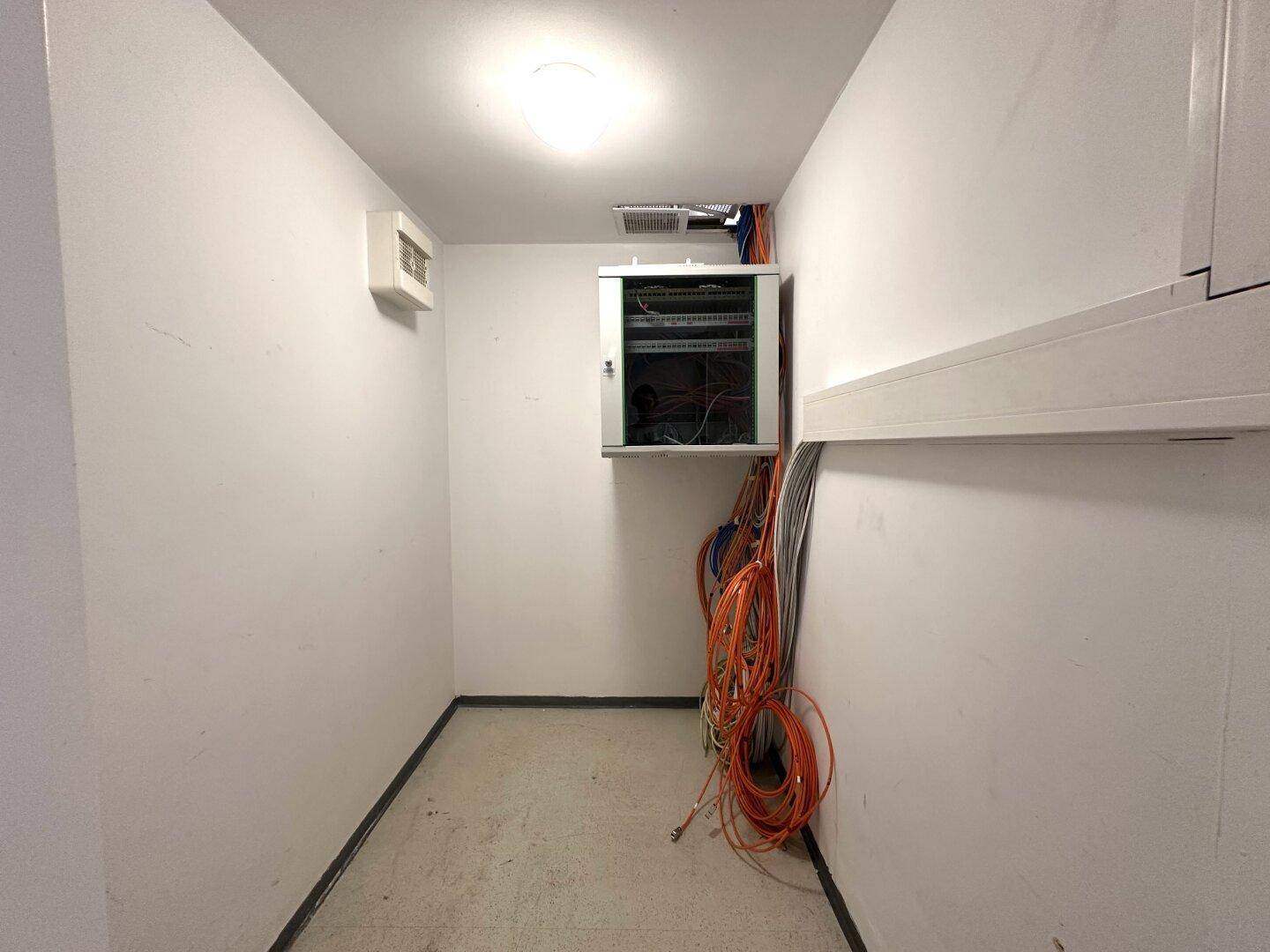 Technikraum mit sichtbaren Kabeln, Netzwerkschrank und einfachem Betonboden.
