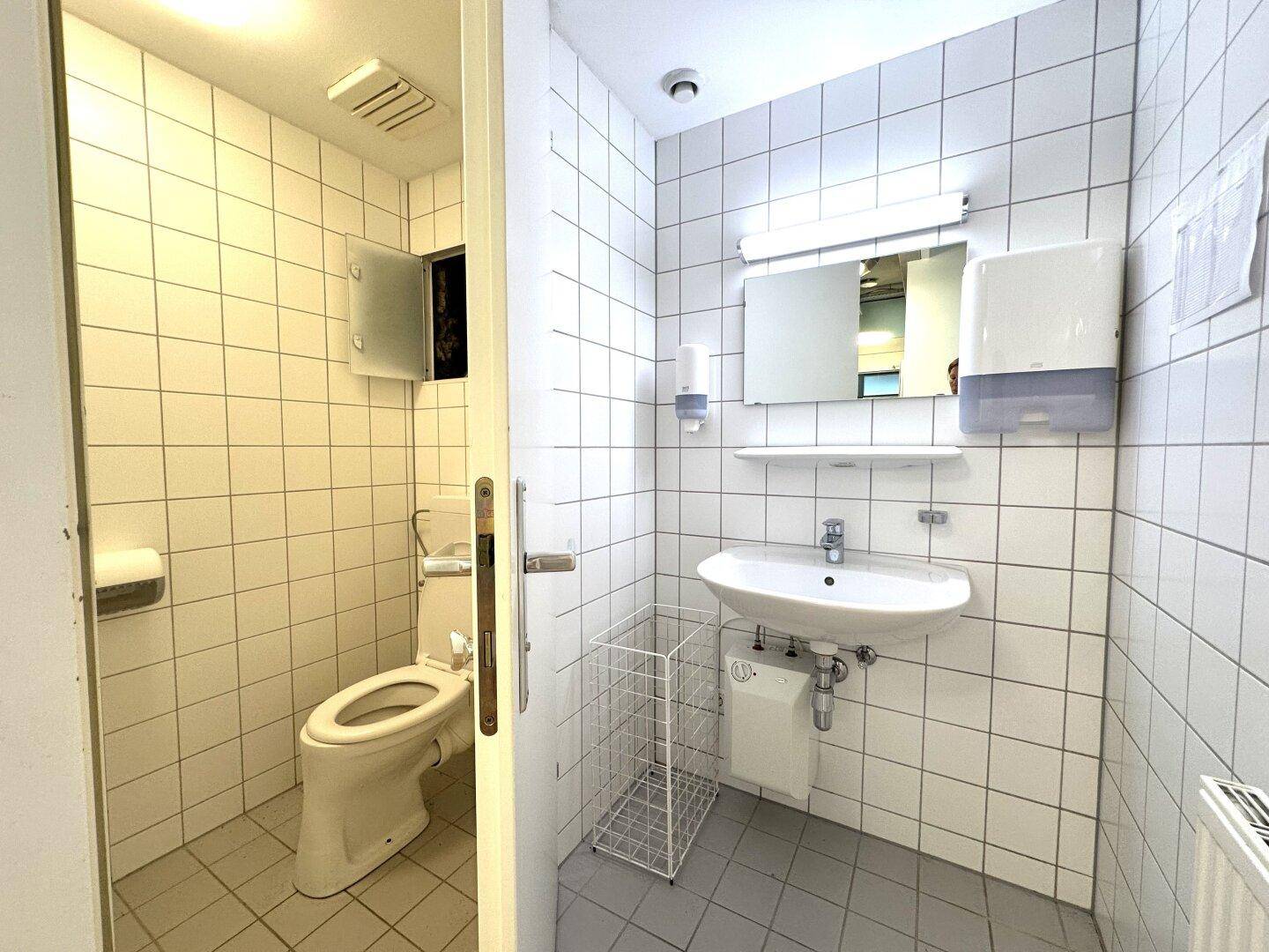 Öffentliche Toilette mit weißen Fliesen, Waschbecken, Spiegel und Haltegriffen an der Toilette.