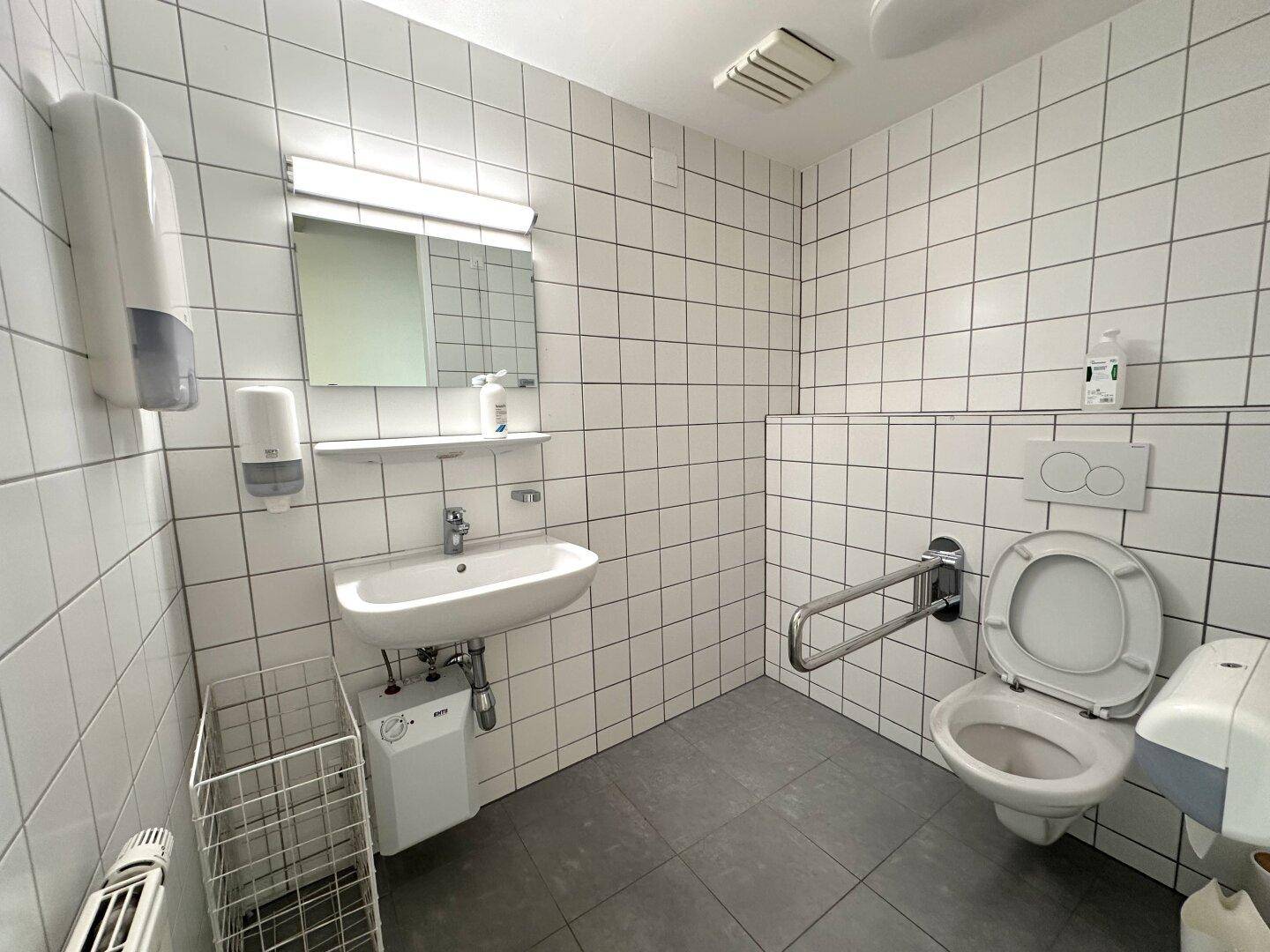 Barrierefreie Toilette mit weißen Fliesen, Waschbecken und Haltegriffen neben der Toilette.