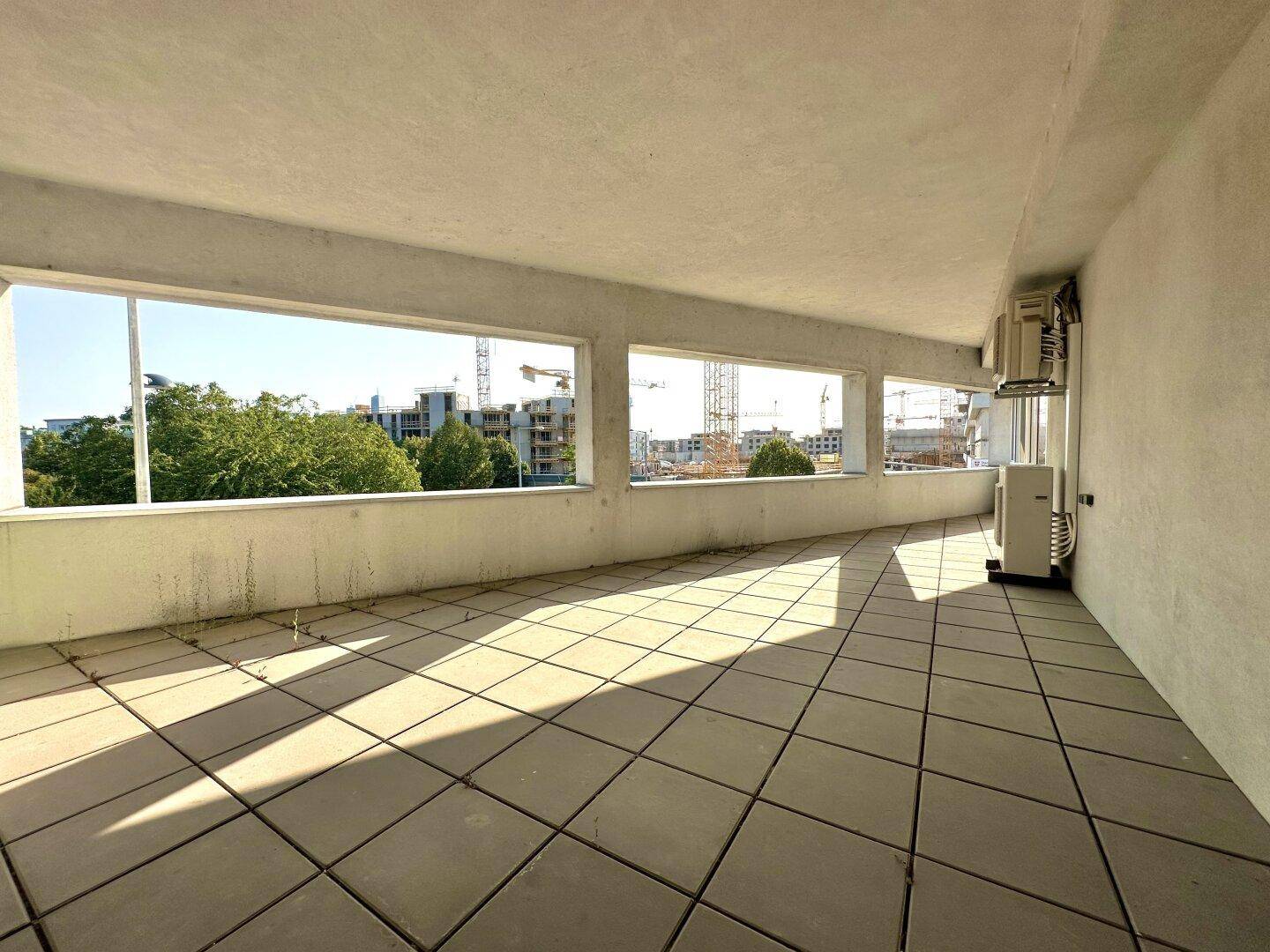 Geräumige Terrasse mit Fliesenboden und Blick auf Baustellen und grüne Bäume in der Ferne.