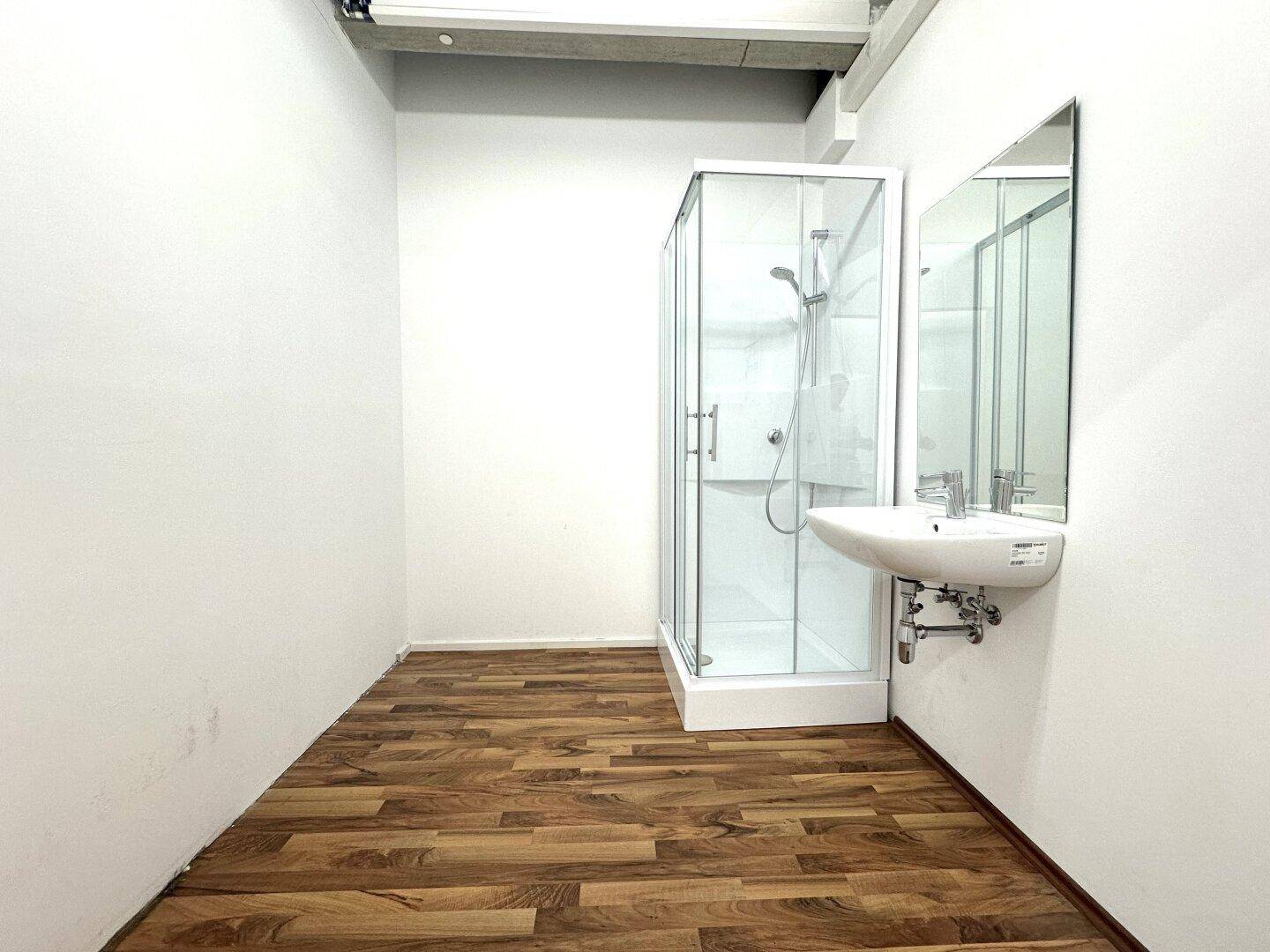 Kleines Badezimmer mit weiß gestrichenen Wänden, Duschkabine und Waschbecken auf Laminatboden.