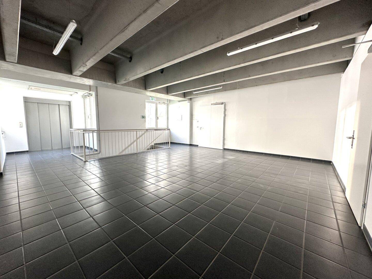 Großer, leerer Gewerberaum mit dunklen Fliesen, Sichtbetondecke und Aufzug im Hintergrund.