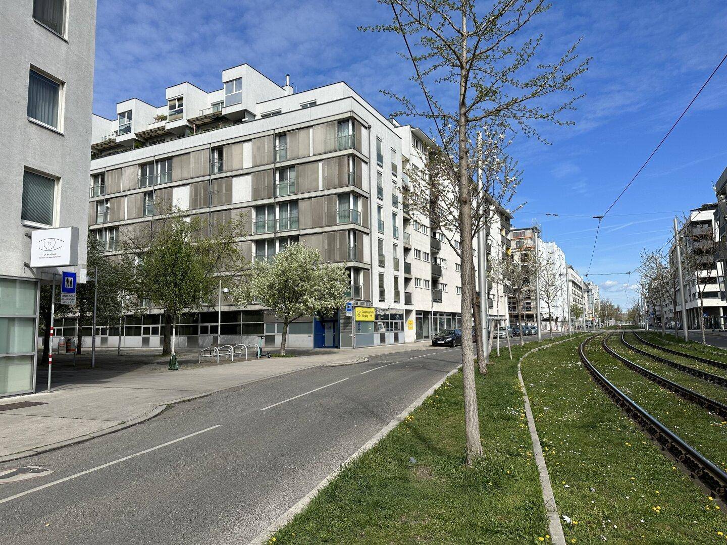 Modernes Gebäude an einer Straße mit Straßenbahnschienen und Bäumen unter blauem Himmel.