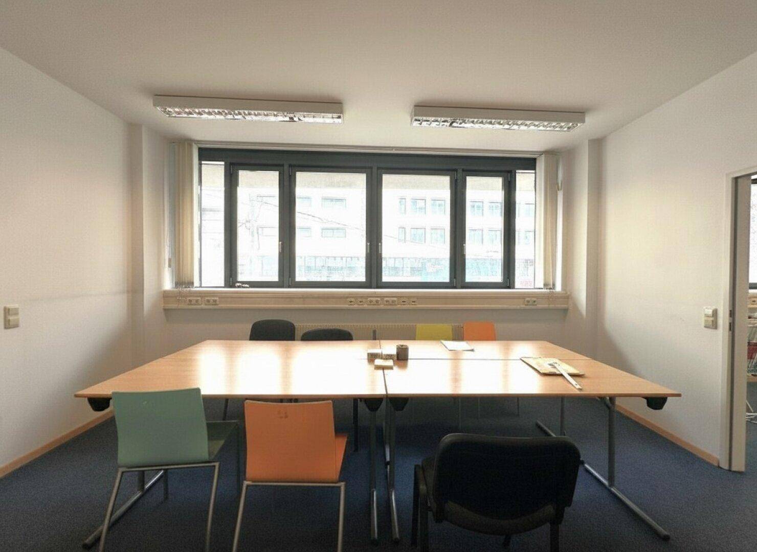 Besprechungsraum mit großem Holztisch, bunten Stühlen und Teppichboden, ideal für Teamtreffen.
