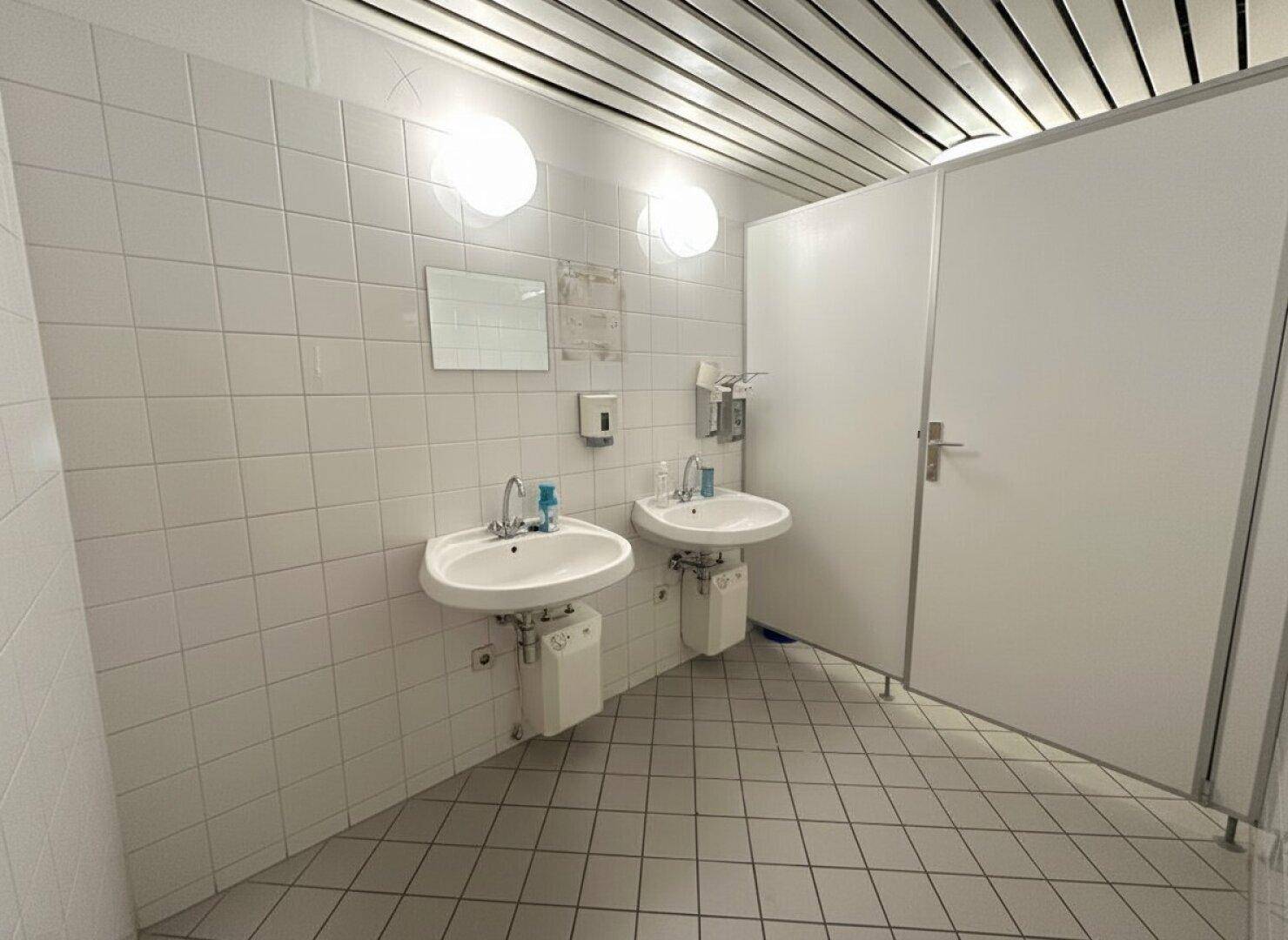 Heller Waschraum mit zwei Waschbecken, weißen Fliesen und einer modernen Deckenverkleidung.