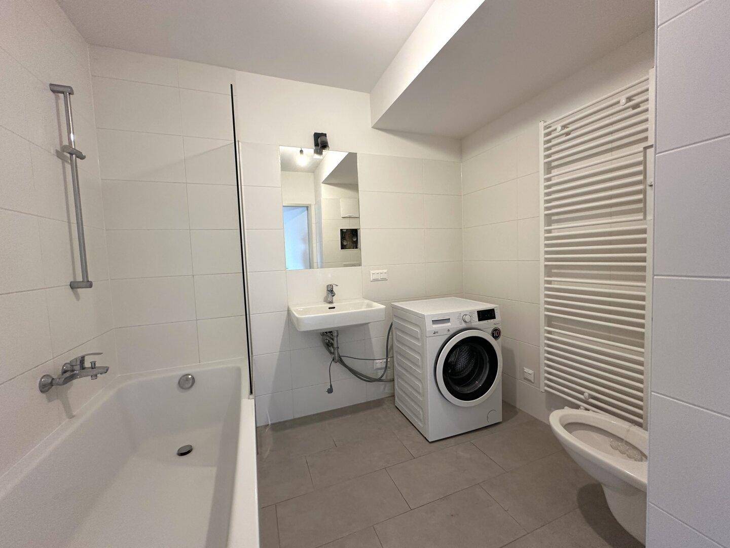 Modernes Badezimmer mit Badewanne, Waschbecken, WC und Waschmaschine, hell gefliest.