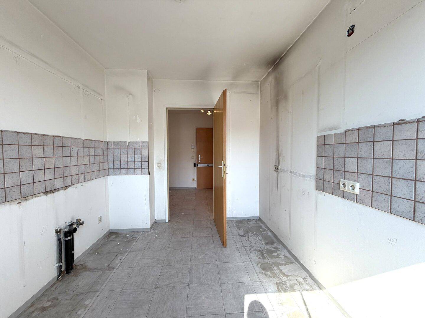 Leere Küche mit Fliesenboden und teilweiser Wandfliesenverkleidung, mit Zugang zu einem weiteren Raum.