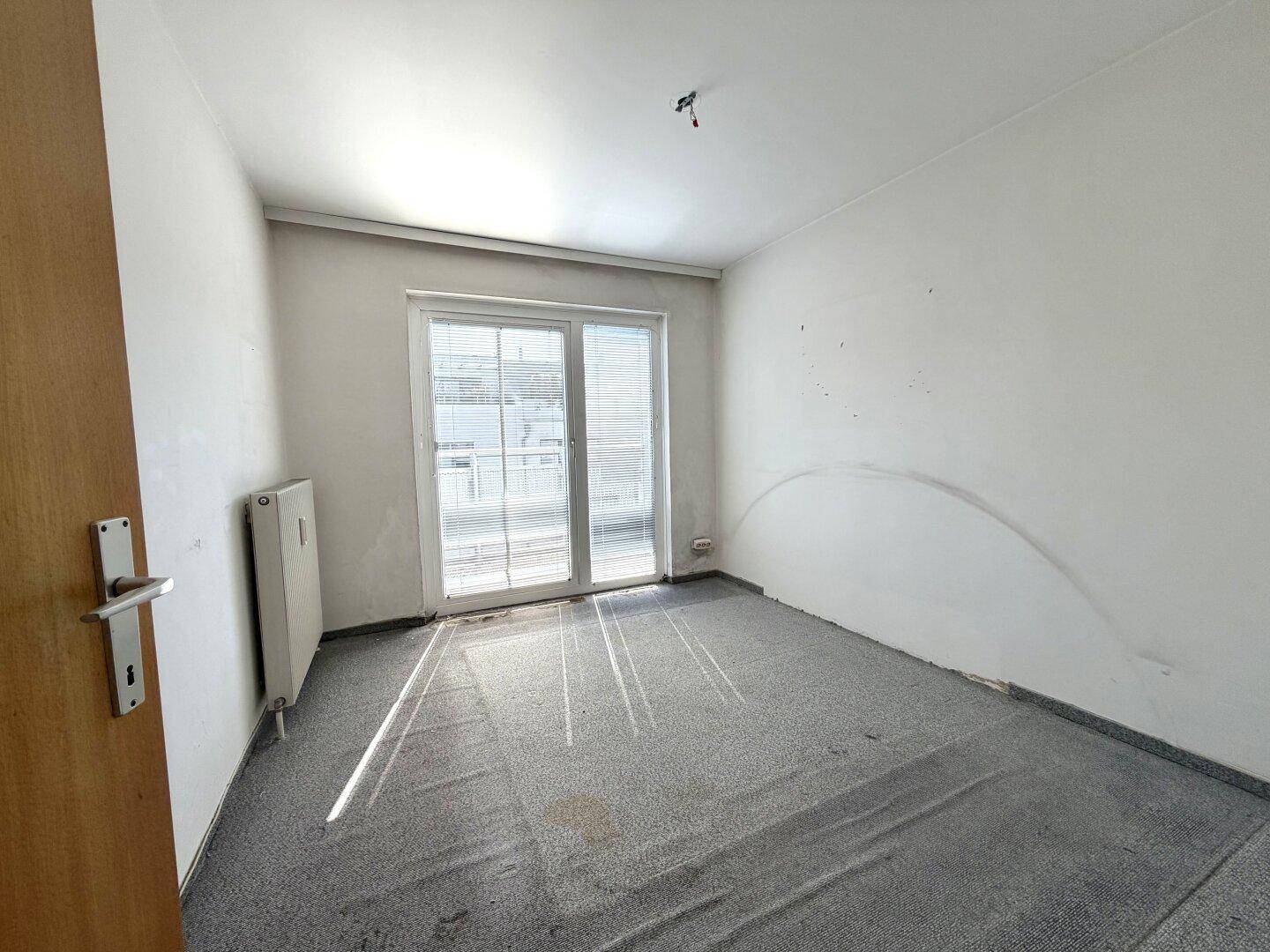 Helles Schlafzimmer mit Teppichboden und großem Fenster, das viel Tageslicht hereinlässt.