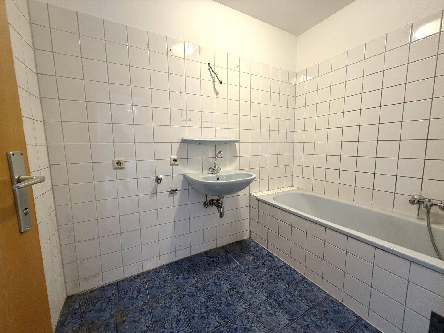 Badezimmer mit Badewanne, Waschbecken und blauen Bodenfliesen, renovierungsbedürftig.