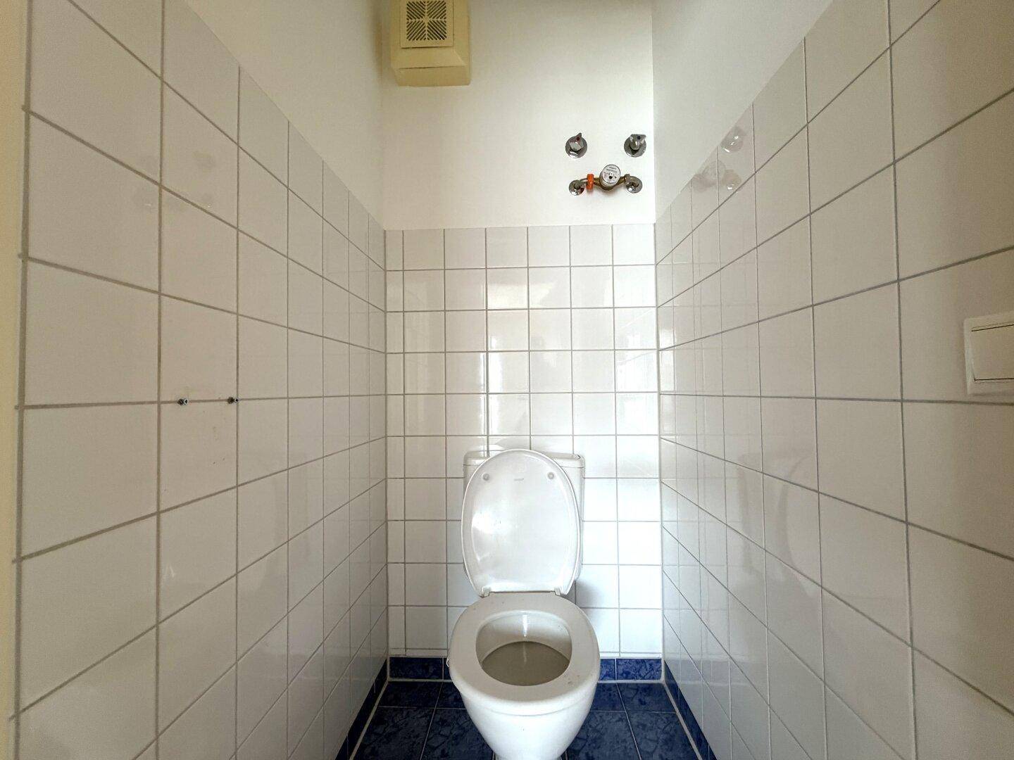 Separate Toilette mit weißen Wandfliesen und blauen Bodenfliesen, mit Abluftventilator.