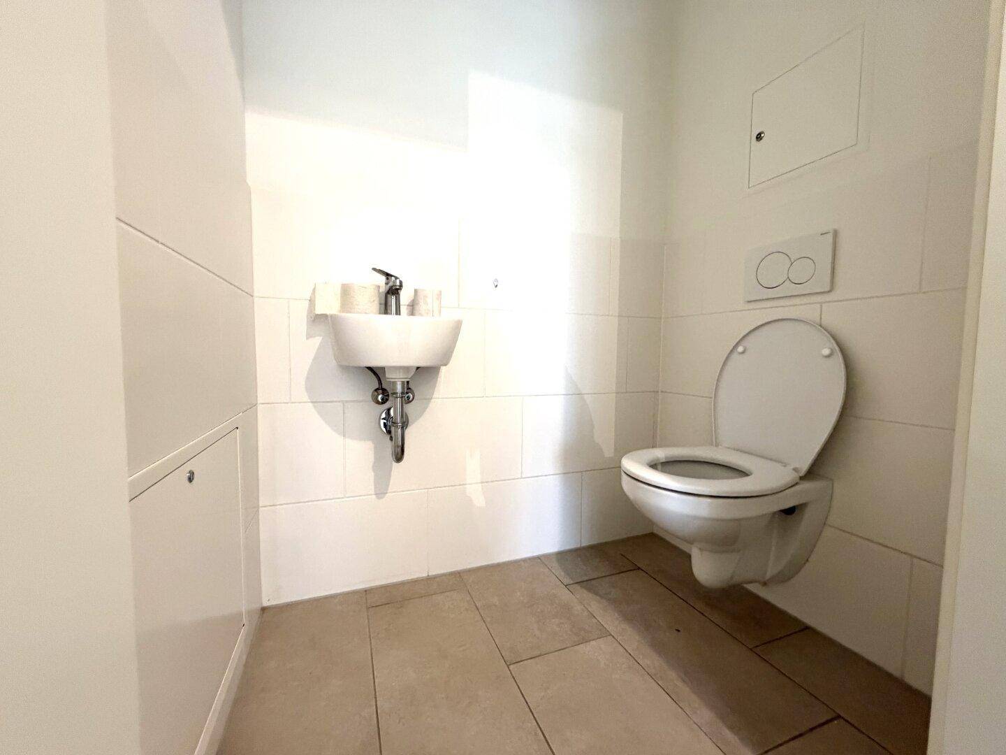 Separates WC mit wandhängender Toilette, kleinem Waschbecken und hellen Fliesen.