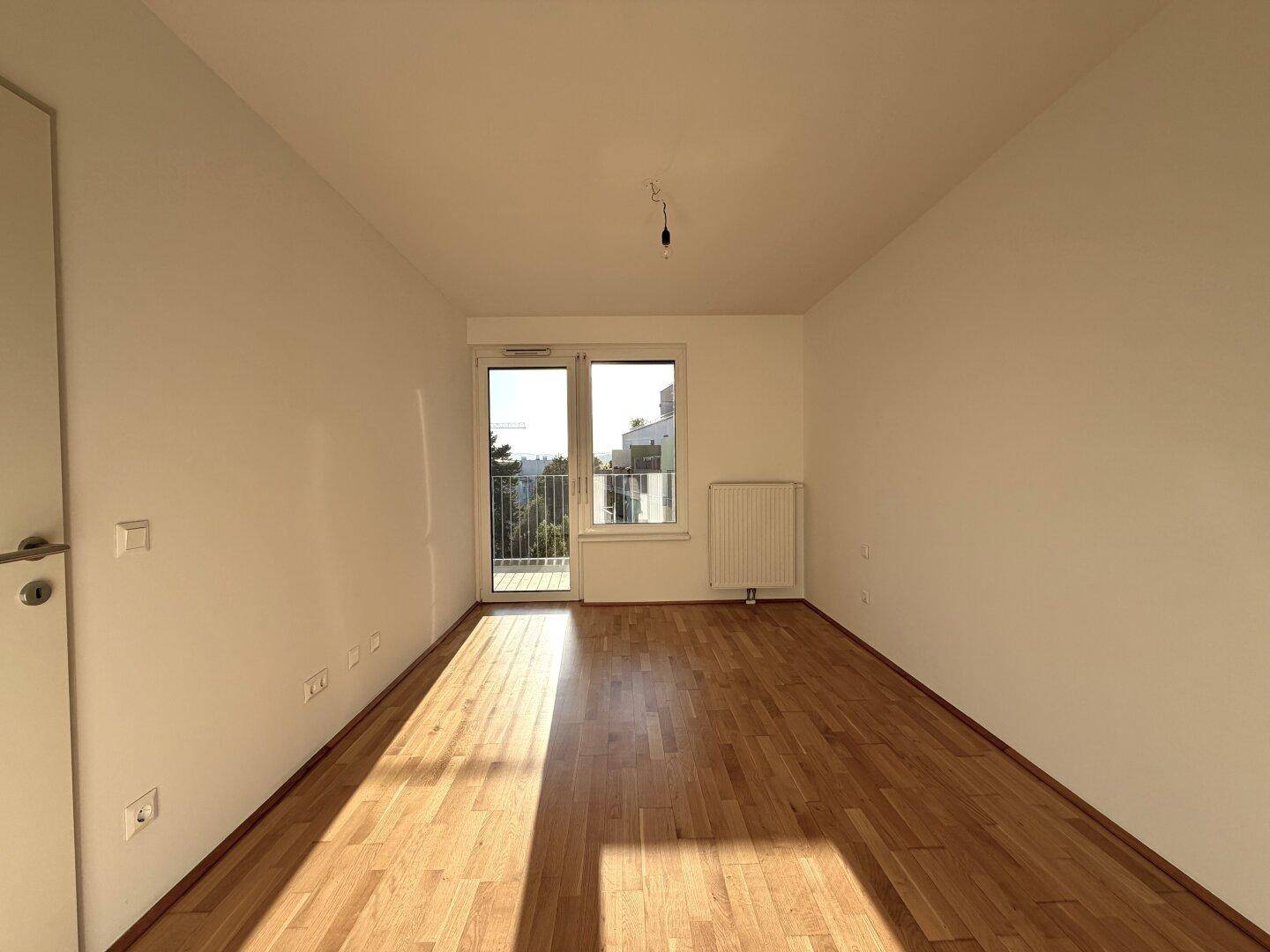 Leeres Zimmer mit Holzboden, großem Fenster und Zugang zu einem Außenbereich.