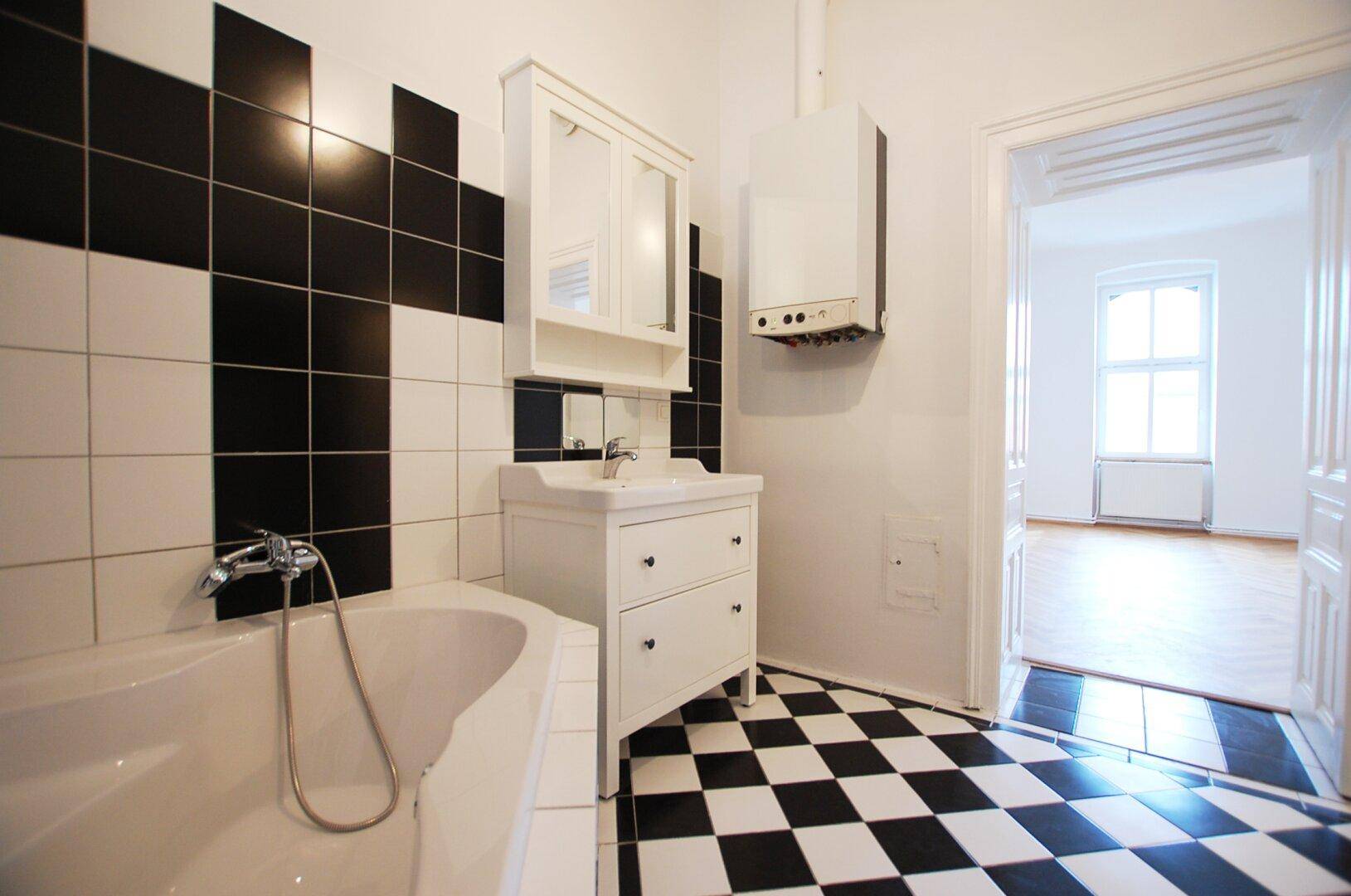 Badezimmer mit schwarz-weißen Fliesen, Badewanne und Blick in den angrenzenden Wohnbereich.