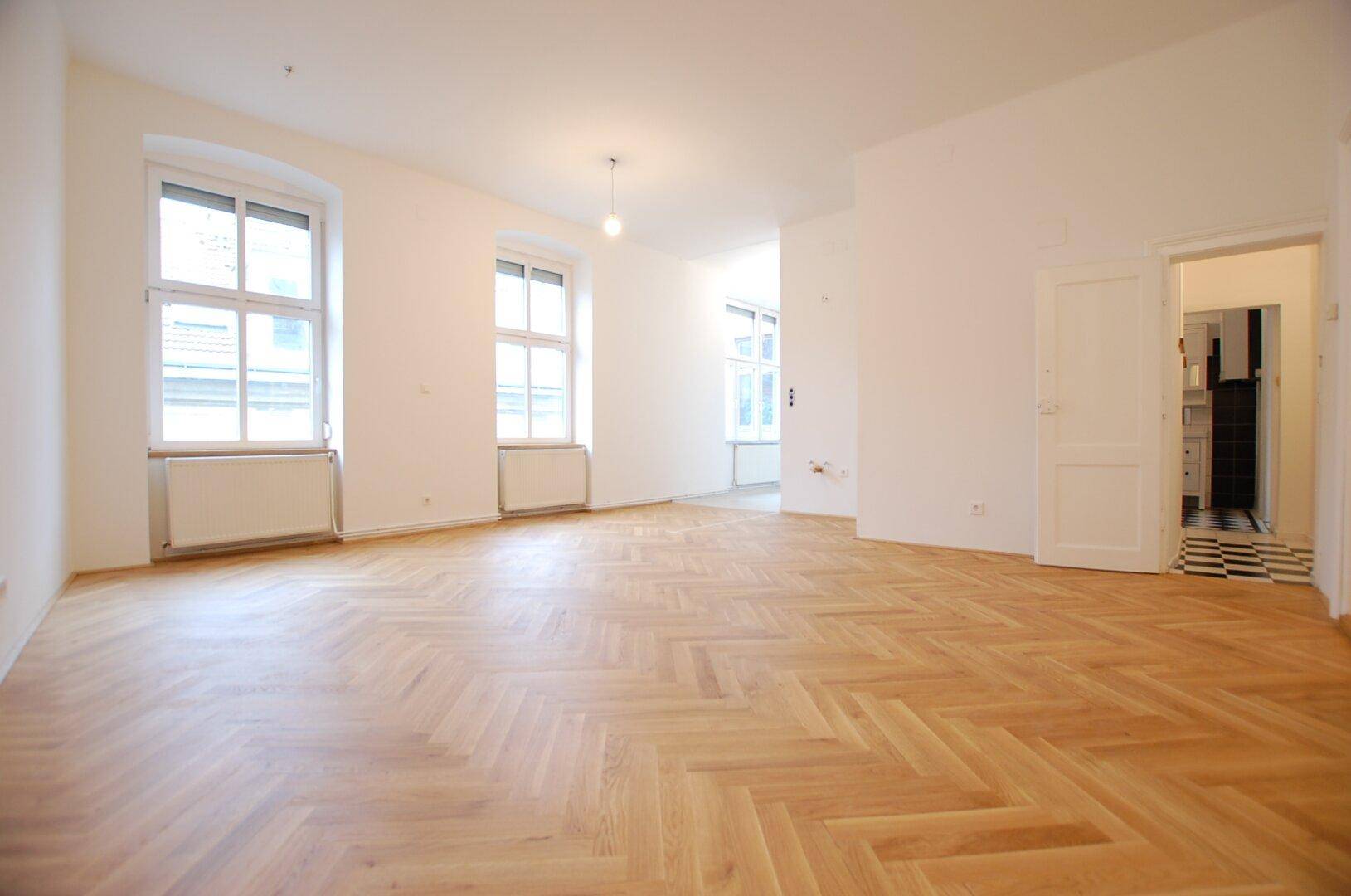 Geräumiges Wohnzimmer mit Fischgrätparkett, hohen Fenstern und viel Tageslicht.
