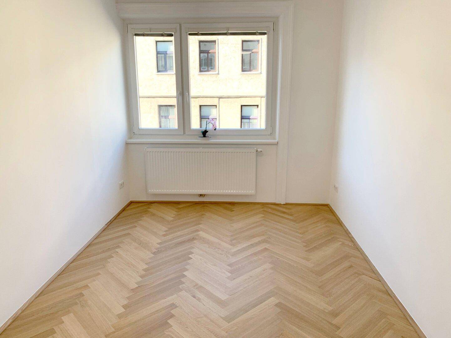 Helles, leeres Zimmer mit Fischgrätparkett und Blick auf die umliegenden Gebäude.