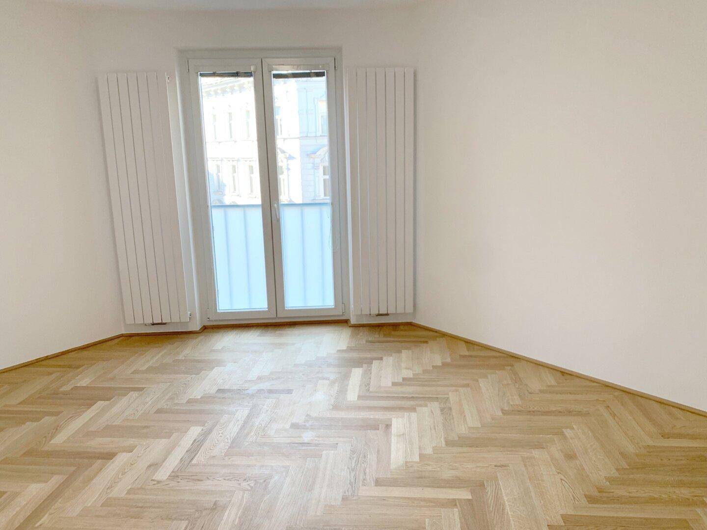 Lichtdurchflutetes Zimmer mit Fischgrätparkett und Zugang zu einem französischen Balkon.