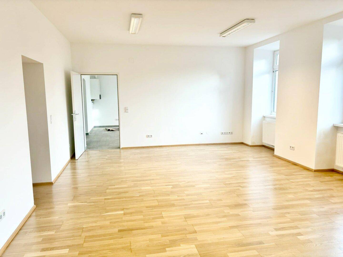 Geräumiger, heller Raum mit Holzparkettboden und einer offenen Tür zu einem weiteren Bereich.