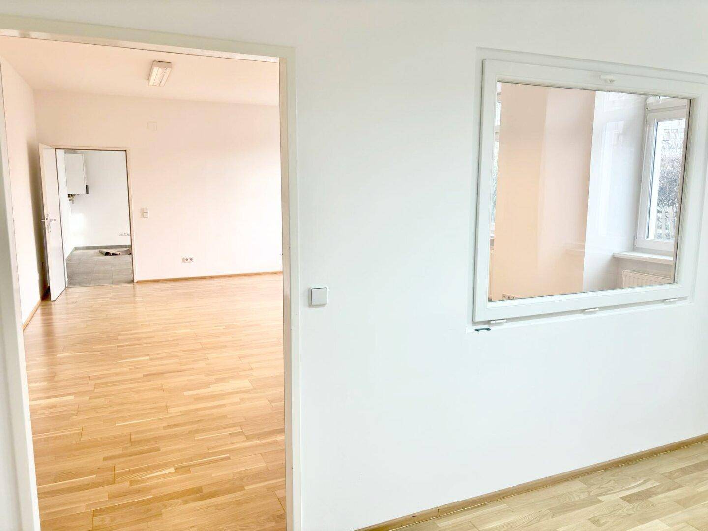 Heller Raum mit Holzparkettboden, einem internen Fenster und einer offenen Tür zu einem anderen Bereich.