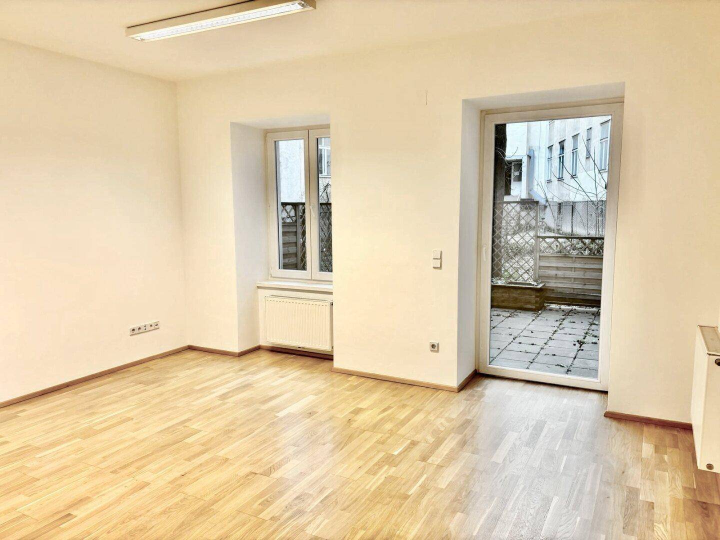 Heller Raum mit Holzparkettboden und einer großen Glastür, die zu einer Außenterrasse führt.