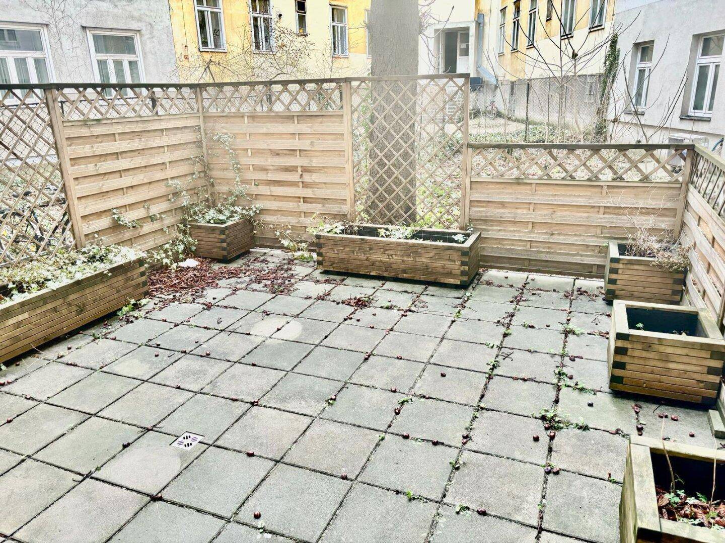 Gepflasterte Außenterrasse mit Holzzäunen und Pflanzkübeln, umgeben von Gebäuden.