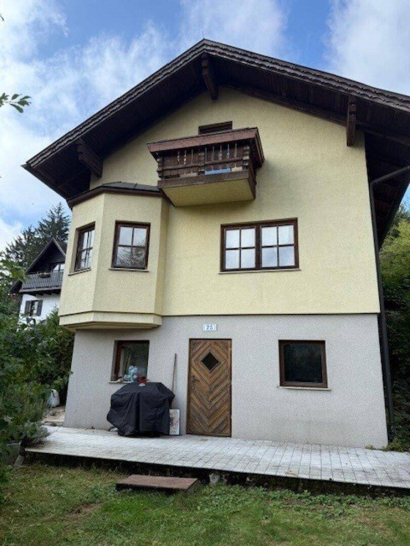 Fassade eines traditionellen Hauses mit Holzbalkon und gelber Putzfassade, umgeben von viel Grün.