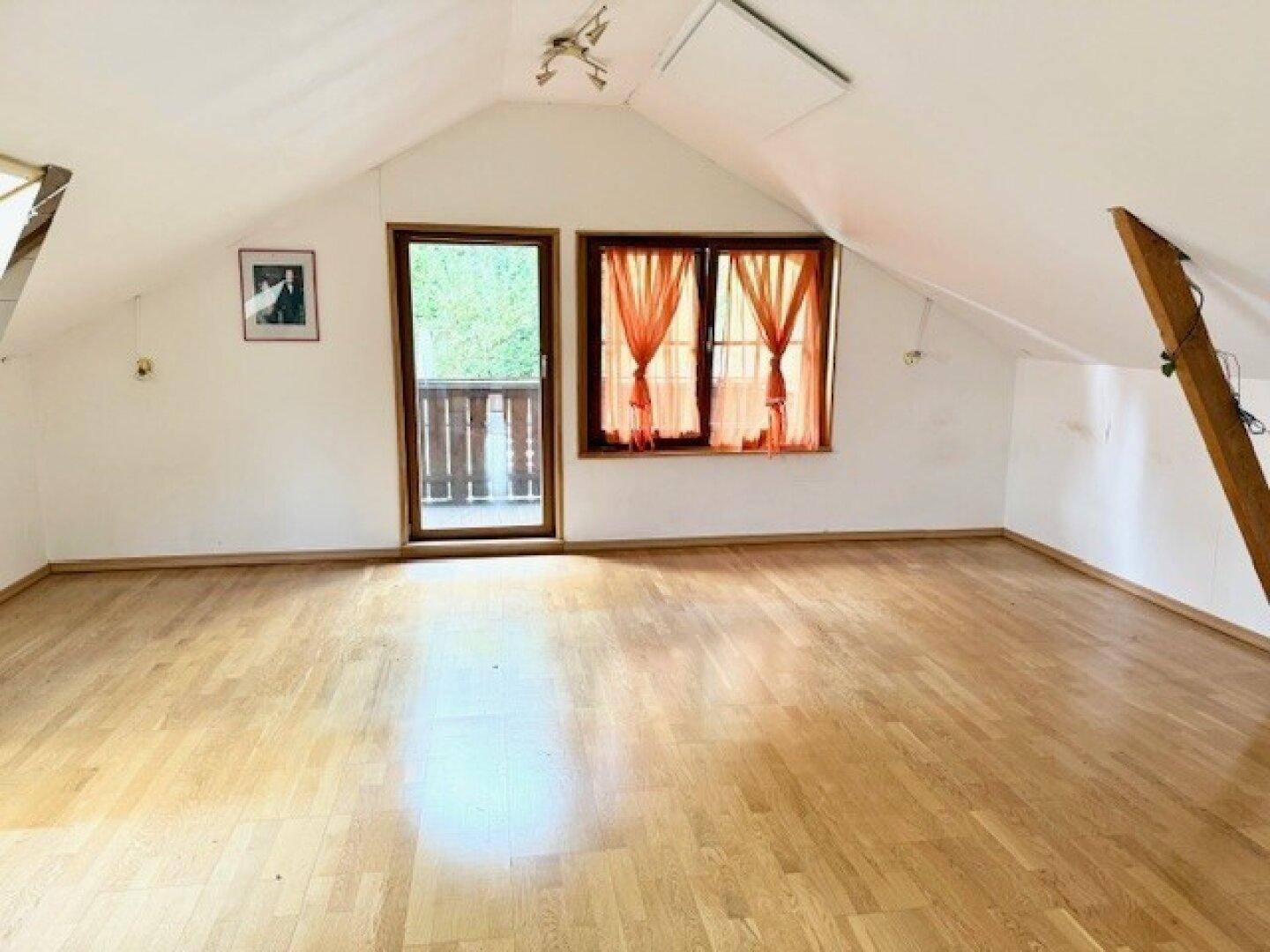 Helles Dachzimmer mit Holzboden, schrägen Decken und Zugang zu einem kleinen Balkon.