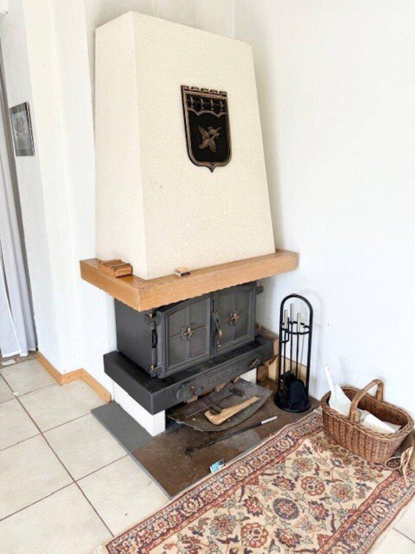 Gemütlicher Kaminofen mit Holzablage und traditionellem Teppich in einem hellen Raum.