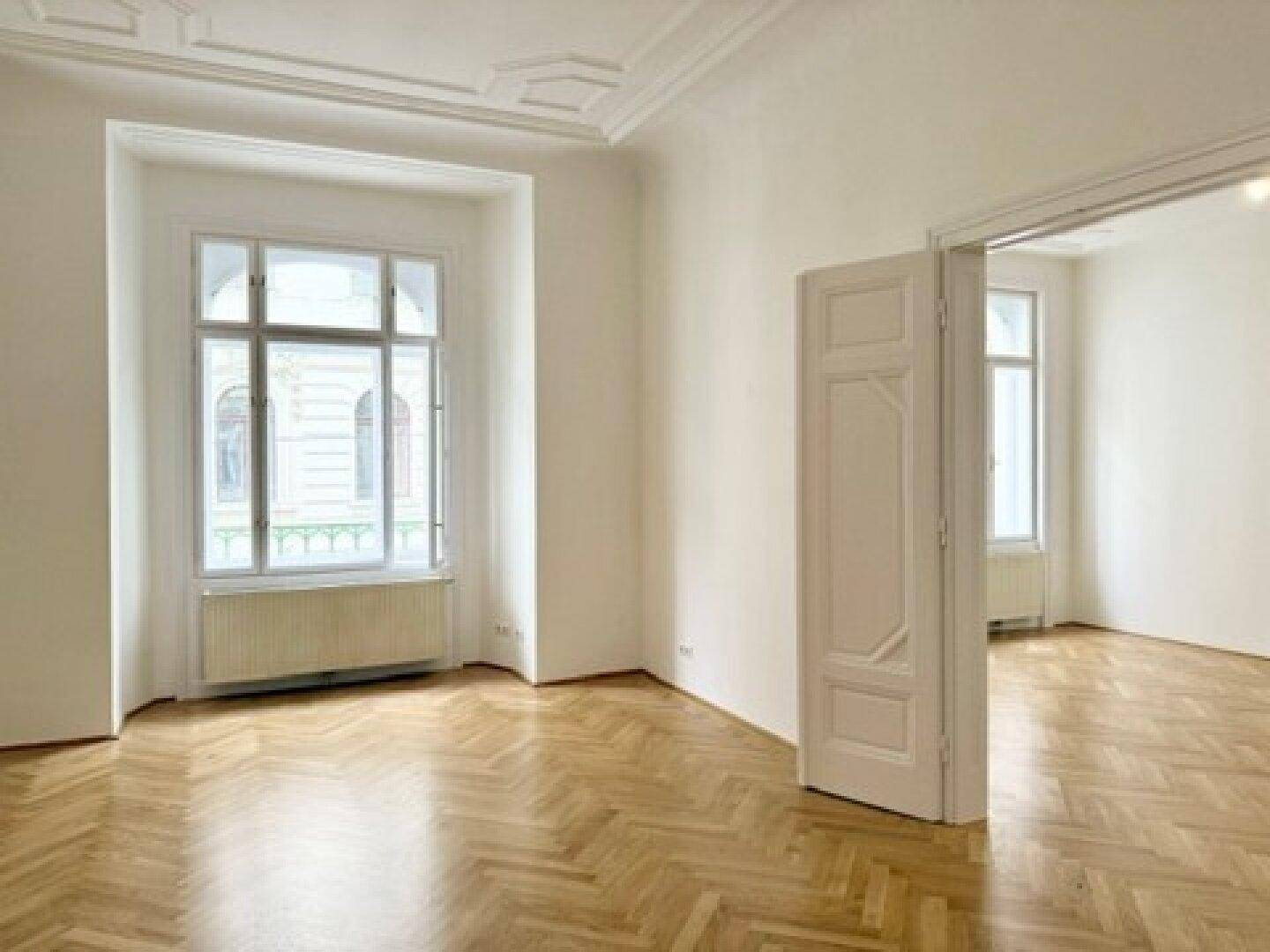 Helles Zimmer mit Fischgrätparkett, hohen Decken und Stuckverzierungen, verbunden durch eine offene Tür.