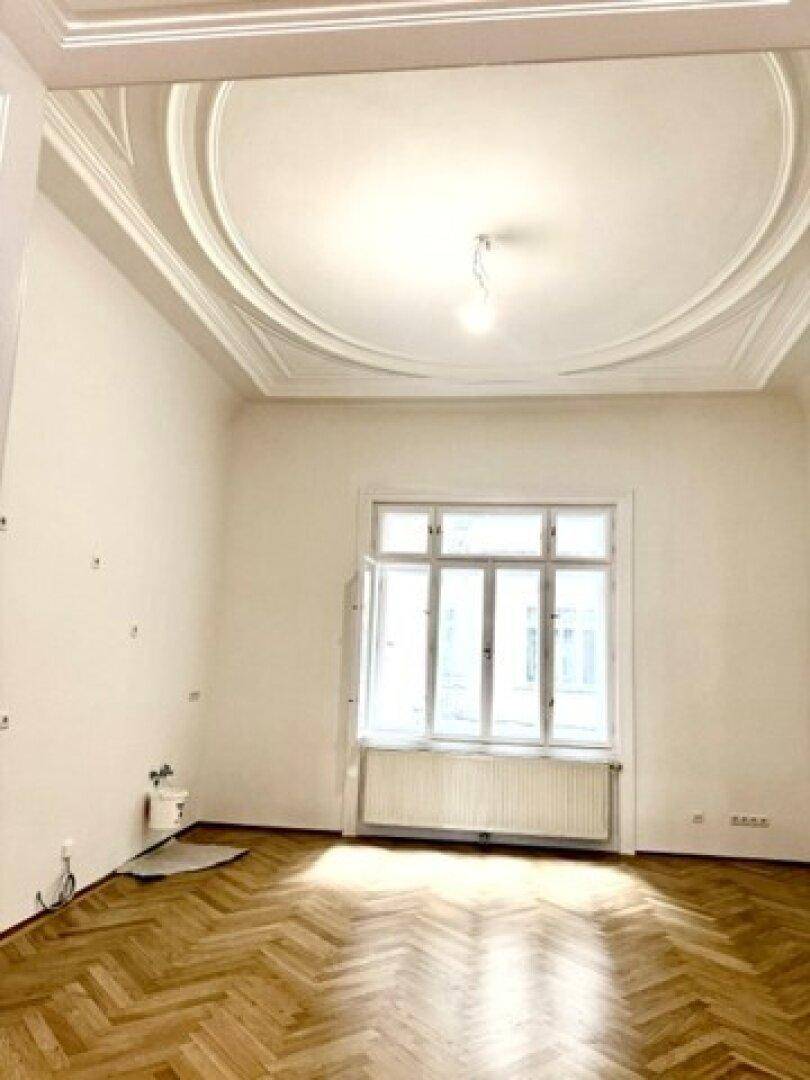 Geräumiges Zimmer mit Fischgrätparkett, hohen Decken und aufwendiger Stuckdecke, gut beleuchtet.