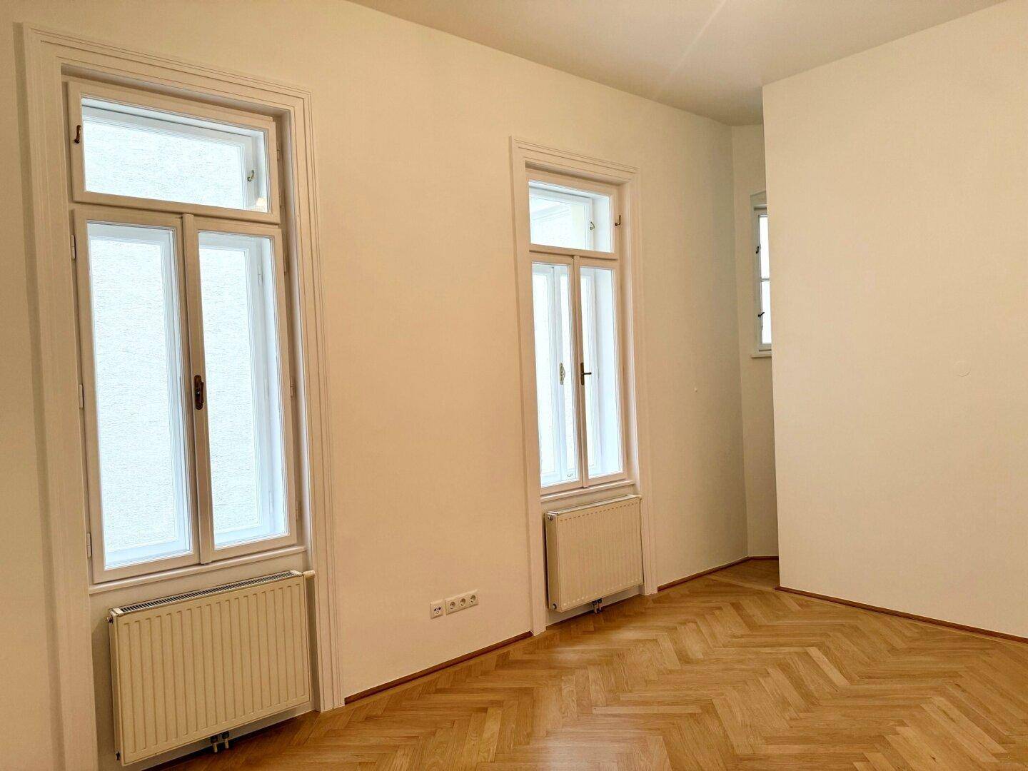 Heller Raum mit Fischgrätparkett und hohen Fenstern, ausgestattet mit Heizkörpern.