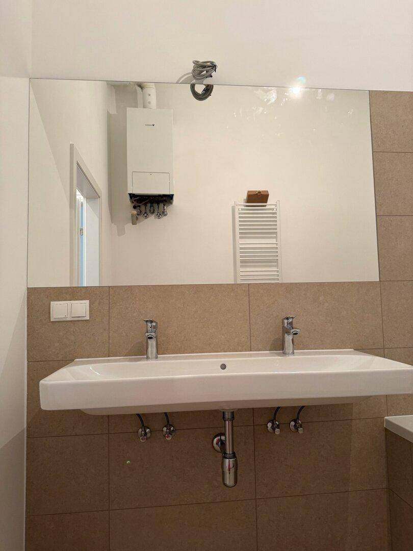 Badezimmer mit Doppelwaschbecken, großem Spiegel und sichtbarem Warmwasserbereiter.
