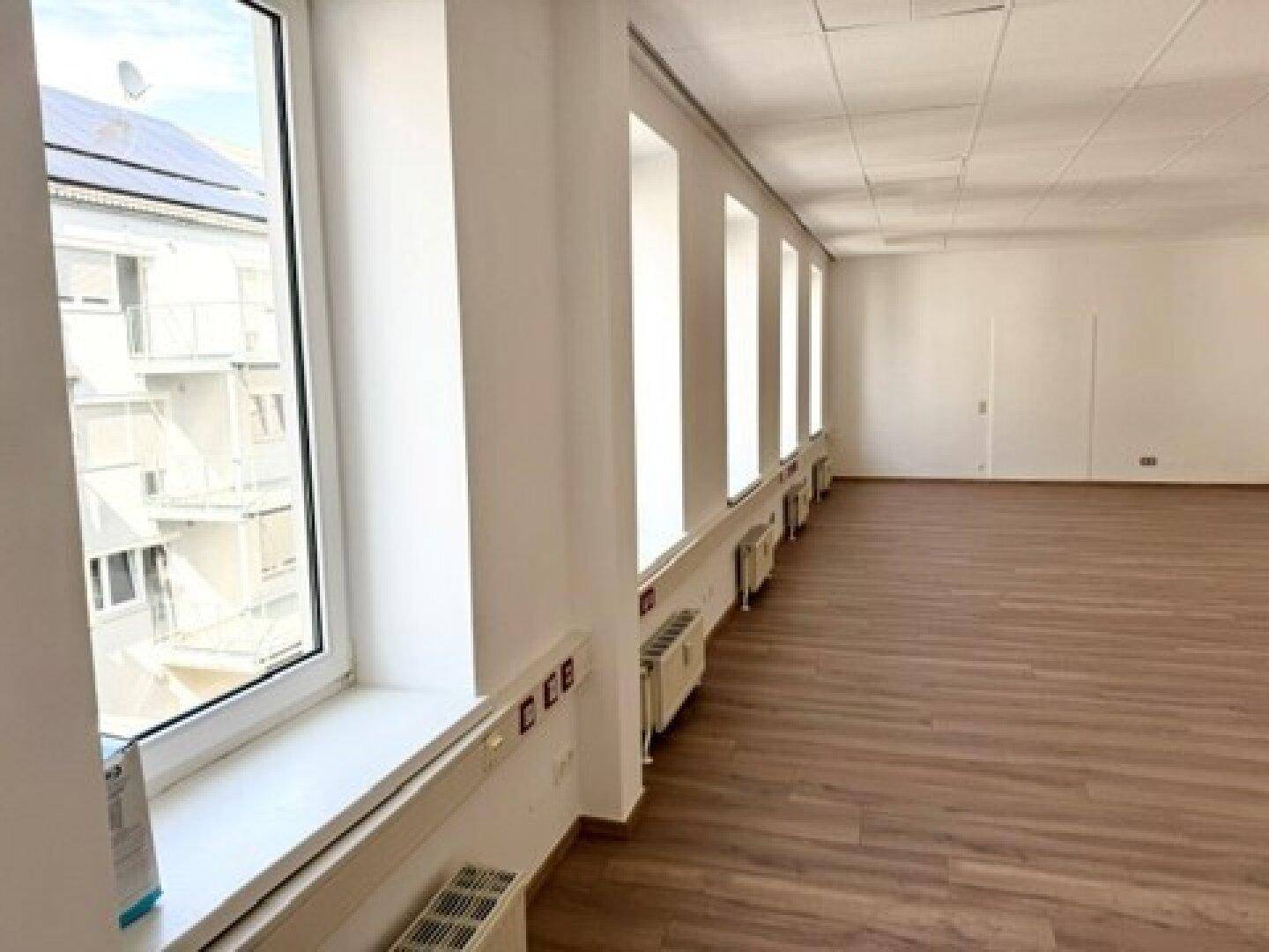 Langer, heller Büroraum mit vielen Fenstern und Blick auf ein Gebäude mit Solaranlage.