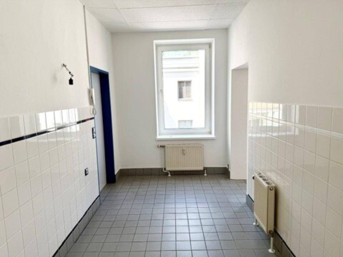 Heller Flur mit weißen Fliesen an den Wänden, grauem Fliesenboden und einem Fenster.
