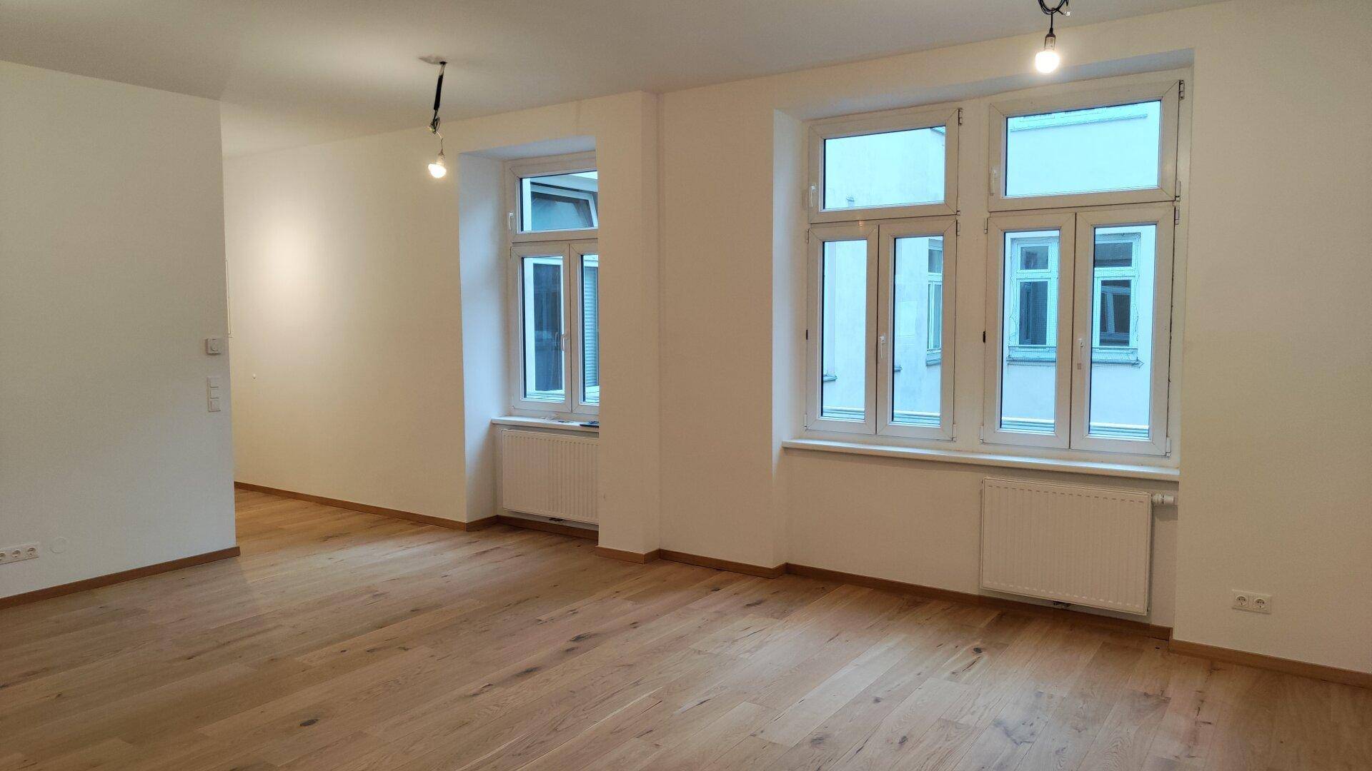 Heller Wohnraum mit großen Fenstern, Holzparkettboden und Heizkörpern unter den Fenstern.