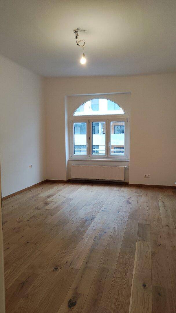 Leerer Raum mit hellem Holzparkettboden und einem großen Bogenfenster.